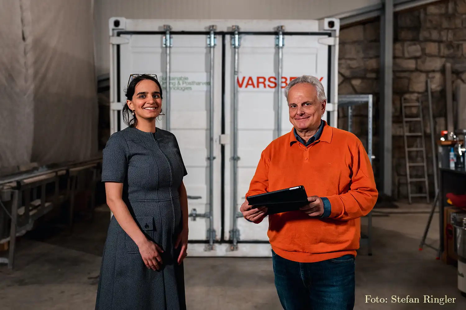 Eine Frau im dunklen Kleid und ein Mann mit einem orangenen Pullover stehen vor einem Container. Der Mann hält ein Tablet in der Hand.